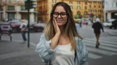 Yuvarlak gözlüklü ve mavi gömlekli genç İspanyol kadın, kalabalık şehir yaya geçidinde elleri göğsünde gülümsüyor; minnettarlık sükuneti.