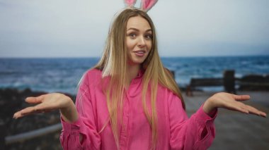 Tavşan kulaklı kadın sahilde yürürken gülümsüyor. Üzerinde pembe bir tişört ve deniz arkaplanı var. Açık havada eğlenceli ve neşeli bir atmosfer yaratıyor..