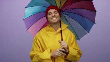 Renkli şemsiyelerin altında gülümseyen genç adam mor arka planda sarı yağmurluk giyiyor..