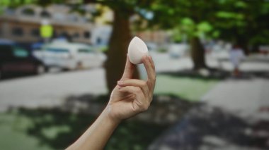 Güneşli bir şehir caddesinde arka planda bulanık arabalar ve ağaçlar olan küçük beyaz bir nesne tutan adam, açık hava ortamını gözler önüne seriyor..