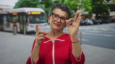 Arka planda bulanık bir otobüsle şehir caddesinde dikilirken işaret eden kırmızı ceketli yaşlı kadın..
