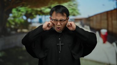 Siyah cüppeli adam şehir sokaklarında kulaklarını kapatıyor, şehir ortamında gürültü karışıklığını vurguluyor..