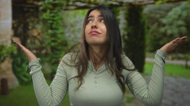 Parkta kafası karışmış, çevresi yeşillik ve açık hava manzarasıyla çevrili, günlük kıyafetler giyen, belirsizlik ve doğada tefekkür taşıyan genç İspanyol kadın..