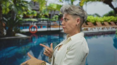 Etrafta ahşap saat, palmiye ağaçları ve şezlonglar olan bir kadın, havuz kenarını işaret ediyor..