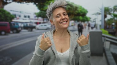 Gri kapüşonlu kadın, bulanık yayalar arasında şehir merkezinde el ele işaret ediyor; davet.