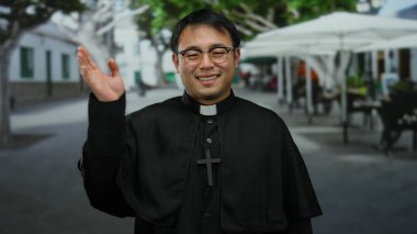 Gözlüklü ve haçlı siyah cüppeli gülümseyen rahip şehir caddesinde ağaçların ve açık hava kafelerin bulanık arka planıyla duruyor..