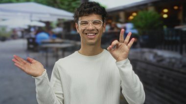 Gözlüklü genç adam avuç içini açık tutuyor ve sokak kafesi terasında boş bir yeri işaret ediyor..