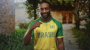 Adam Brezilya formasını ve bayrak yamasını her iki eliyle bina girişinde gösterirken gülümsüyor ve diş tellerini gösteriyor; takım gururu.
