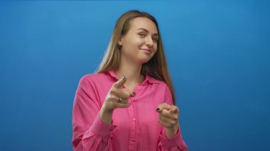 Mavi arka planda gülümseyen ve kameraya işaret eden bir kadın, izole edilmiş bir duvarın üzerinde canlı pembe bir tişört giyip pozitiflik ve kendine güven gösteriyor..