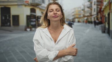 Gözleri kapalı ve kolları çapraz beyaz bir şehir caddesinde kendini kucaklayan kadın, rahat duruş; dinginlik.
