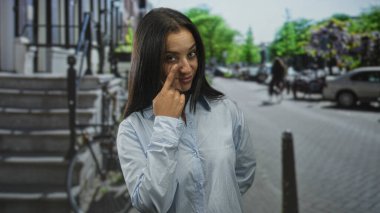 Kadın, açık mavi gömlek ve ince bir gülümseme giyen park halindeki arabaların ve bisikletin yanındaki bir caddede göz göze gelir..