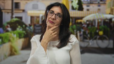 Gözlüklü ve beyaz bluz giyen orta yaşlı bir kadın, kentin işlek caddelerindeki kafede el pençe üstüne el hareketleriyle bisikletler görünür; iç gözlem tefekkür.