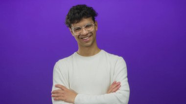 Gözlüklü İspanyol genç adam ve kolları mor stüdyo fotoğrafında çaprazlanmış beyaz süveter, kameraya gülümsüyor;.
