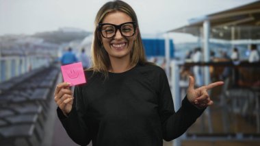 Kadın, korkuluklar ve koltuklar görünür olduğu halde güvertede gülümserken tuttuğu pembe yapışkan notayı işaret ediyor; iyimserlik.