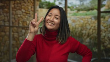 İçerde taş duvar arkası olan barış işaretleri gösteren bir kadın. İyi aydınlatılmış rahat bir odada kırmızı süveter giyiyor. Neşeli ve olumlu duygular ve mutlu bir ifade sergiliyor..