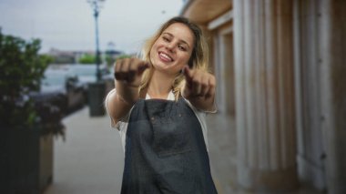 Kadın, kot önlük ve beyaz tişört giyen, doğrudan izleyiciye gülümseyen, neşeli bir davetiye ile şehir caddesinde bir kamerayı işaret ediyor..
