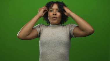 Afrikalı Amerikalı kadın kıvırcık saçlarını yeşil stüdyo ortamında odaklanmış ifadeyle ayarlıyor..