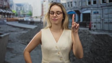 Krem rengi yelek ve yuvarlak gözlüklü bir kadın şehir caddesinde inşaat molozları ve mağazaların yanında işaret parmağını havaya kaldırıyor..