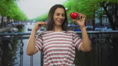 Çizgili tişörtlü genç İspanyol kadın Amsterdam 'da bir sokak köprüsünde kırmızı kalp ve pısırık tutuyor; kalp kırıklığı kaybı.