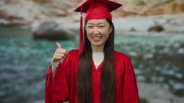 Kırmızı mezuniyet elbisesi giymiş bir kadın deniz kenarındaki açık hava ortamında akademik başarısını kutlarken bulanık bir geçmişle deniz kenarına gülümsüyor..