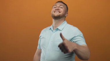 Stüdyoda gülümseyen ve baş parmak hareketi yapan bir adam, turuncu arka planda başparmak ve önkol gösteren bir jest yapıyor..