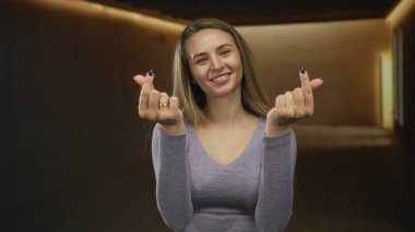 Kapalı alanda gülümseyen, loş bir odada parmaklarını hareket ettiren, mor bir gömlek giyen, arkadaş canlısı, davetkar bir tavırla neşe ve sıcaklık ifade eden, uzun saçlı bir kadın..