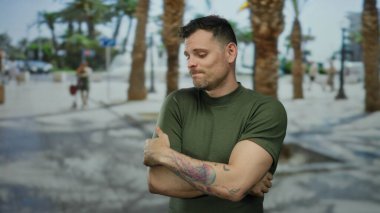 Şehir sokağında palmiye ağaçlarıyla dikilen, yeşil tişörtlü, kolunda dövme olan, düşünceli ve kendini kucaklayan bir adam..