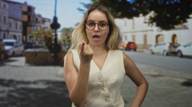 Güneş ışığıyla aydınlatılmış bir şehir caddesinde el hareketi gösteren ön planda gözlük ve kolsuz yelek giyen bir kadın kamerayı işaret ediyor..