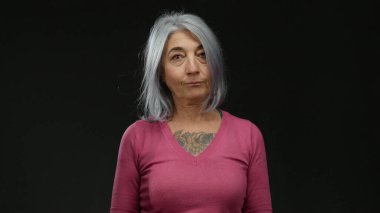Yaşlı, gri saçlı, dövmeli ve pembe kazaklı bir kadın. Stüdyo ortamında derin düşüncelere daldığını ifade eden siyah bir arkaplan karşısında izole edilmiş..