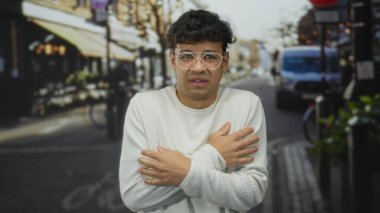 Beyaz kazaklı genç İspanyol adam bulutlu gökyüzü altındaki kafelerle dolu kalabalık bir şehir sokağında ellerini ovuşturuyor ve titriyor;.