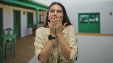 Verandada elleriyle ağzını kapatan kadın, gülümseyen ve avuçlarını yüzüne doğru kaldıran; sevinç sürprizi.
