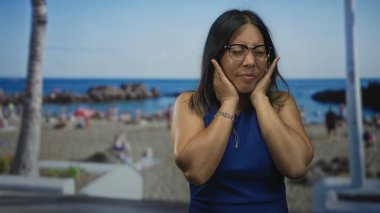 Mavi kolsuz bluzlu bir kadın stüdyo sahili arka planında gözleri kapalı, yanaklarına el ele tutuşuyor, gözlük takıyor; dinginlik.