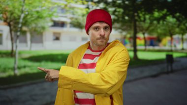 Sarı ceketli genç adam parkta jest yapıyor. Arka planda yeşil ağaçlar var.