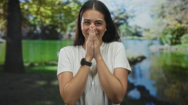 Elleri duada birbirine basılı gülümseyen, beyaz tişört giyen ve ormanda gölün kenarında duran kadın, nazikçe minnettarlık..