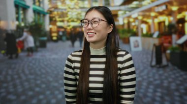 Şehir caddesinde gülümseyen gözlüklü bir kadın. Bulanık şehir arkaplanının ortasında canlı ışıklarla şehir atmosferini yakalıyor ve hareketli yaya aktivitelerini yakalıyor.