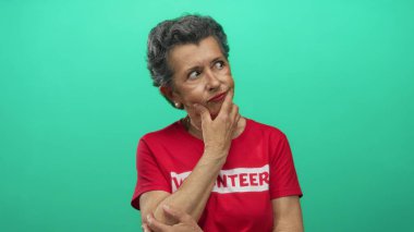 Gönüllü tişört giyen gri saçlı yaşlı bir kadın canlı yeşil bir arka plana karşı düşünceli bir şekilde duruyor, yansıma ve toplum hizmeti duygusunu somutlaştırıyor..