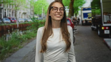 Bir şehir caddesinde park halindeki arabalarla dolu bir kanalda gülümseyen ve el sallayan gözlüklü kadın..