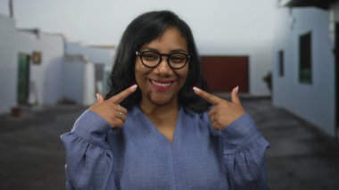 Mavi bluzlu, gözlüklü bir kadın, eski kasaba sokaklarında beyaz bir binanın yanında gülümseyen yanaklarını işaret ediyor..