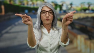 Son sınıf öğrencisi, gri saçlı, parmaklarıyla hayır işareti yapan bir kadın. Sokakta öylesine dikilmiş, vücut dili ve duygularını gözler önüne seriyor..