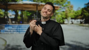 Siyah cüppeli Katolik bir rahip elinde bir haç, açık hava parkı ortamında, güneş ışığı altında huzur ve yansıma simgeliyor..