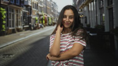 Çizgili gömlekli genç bir kadın, dar bir kaldırım taşı şehir caddesinde elleri kıllı, kameraya gülümsüyor; kaygısız yaz neşesi.