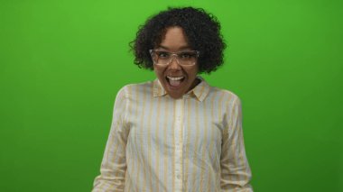 Kıvırcık saçlı, gözlüklü ve çizgili tişörtlü bir kadın yeşil stüdyo resminde gülümsüyor; neşeli iyimserlik.
