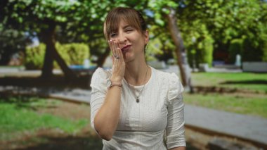 Kadın elinle yanağa dokunuyor ve ormanda avuç içi kaldırıyor park yolunun yanında beyaz bluz ve kol saati giyiyor, düş kırıklığı.