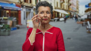 Gri saçlı yaşlı bir kadın şehir arka planına karşı bir jest yapıyor. Gün boyunca açık havada kırmızı bir bluz giyiyor..