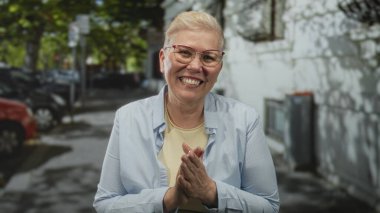 Woman clapping hands on a city street while smiling broadly and wearing glasses and casual shirt; warmth joy.