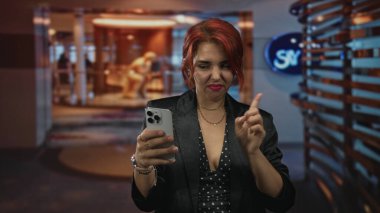 Genç İspanyol kadın elinde akıllı telefonlar ile otel lobisinde siyah ceket ve puantiyeli bluz giyerek ekranı işaret ediyor..