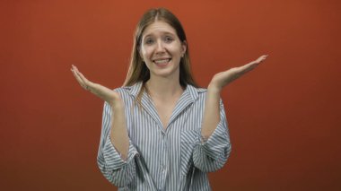 Sarı saçlı, çizgili tişörtlü bir kadın iki elini de stüdyoda tutarak turuncu arka planda belirsizlik yaratıyor..