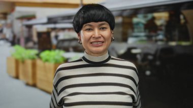 Açık teras restoranında parmaklarıyla bardaklar oluşturan bir kadın, çizgili bir kazakla, arkasında bitkiler ve yapılar olan sıradan bir ortam sergiliyor..