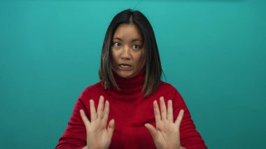 İzole edilmiş yeşil bir duvara karşı sakin olmak için el kol hareketi yapan, kırmızı kazak giyen, sakin bir ifadeye ve genç bir Çinli görünümüne sahip bir kadın..