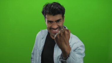 Genç İspanyol adam yeşil stüdyoda sıkıca yumrukluyor kot gömlek ve siyah tişört giyiyor; kararlılık.
